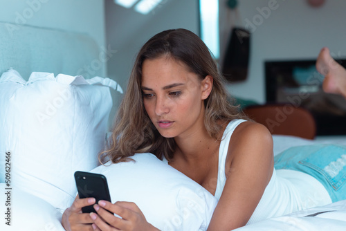 Caucasian young woman using smartphone lying on the bed at home