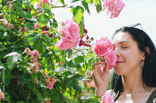 Lifestyle Portrait. Beautiful happy woman walking, relaxing in sunny day at garden with blooming pink roses. smelling flowers. Summer