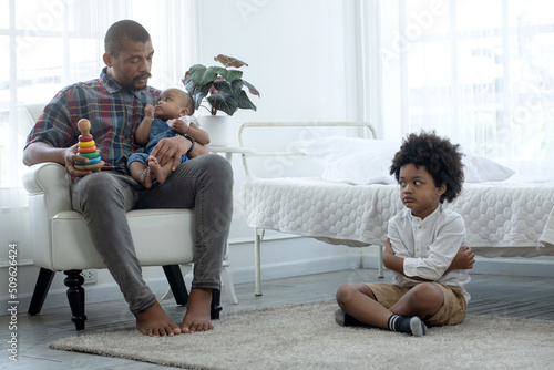 African father holding and playing with little baby girl, child son siting on floor and turned his back and was displeased