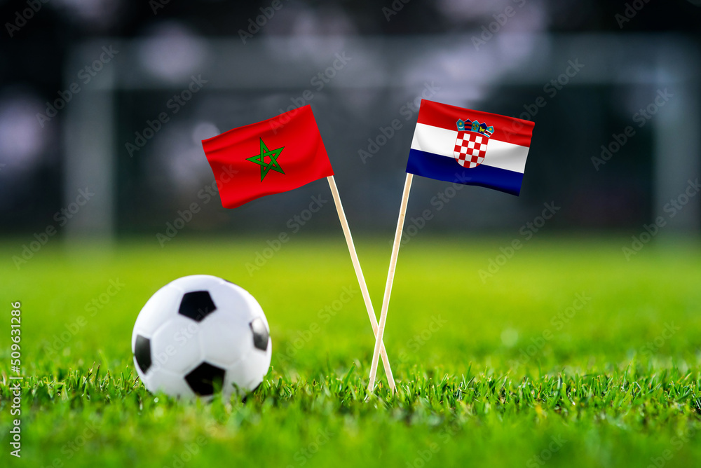 Fototapeta premium Morocco vs. Croatia, Wed, Nov. 23, Al Bayt, Football match wallpaper, Handmade national flags and soccer ball on green grass. Football stadium in background. Black edit space.