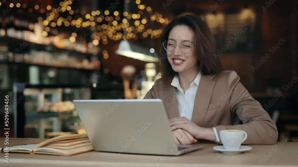 Smiling caucasian brunette in glasses and jacket using modern laptop for video call while sitting at cafe table. Concept of people, business and online conversation. Smiling caucasian brunette in glasses and jacket using modern laptop for video call while sitting at cafe table. Concept of people, business and online conversation.
