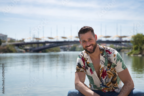 Young and handsome man, blue eyes, perfect smile, beard and Hawaiian shirt, sitting on a pier by the river, very smiling. Concept vacations, party, trips, cruises. © Manuel
