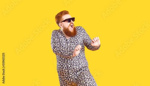 Crazy plus size man in funny pajamas dancing and having fun in fashion studio. Happy bearded fat guy wearing cool sunglasses and comfy leopard PJs singing and dancing isolated on yellow background