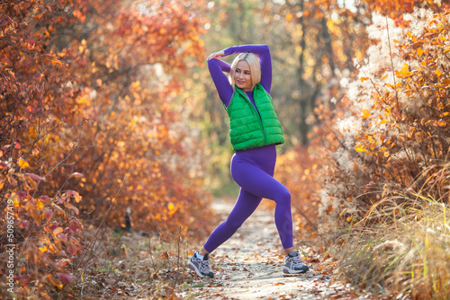 Workout outdoors concept. Young blond athletic woman in sportswear practices aerobics, warming up before training or stretching in the autumn forest. Healthy lifestyle.
