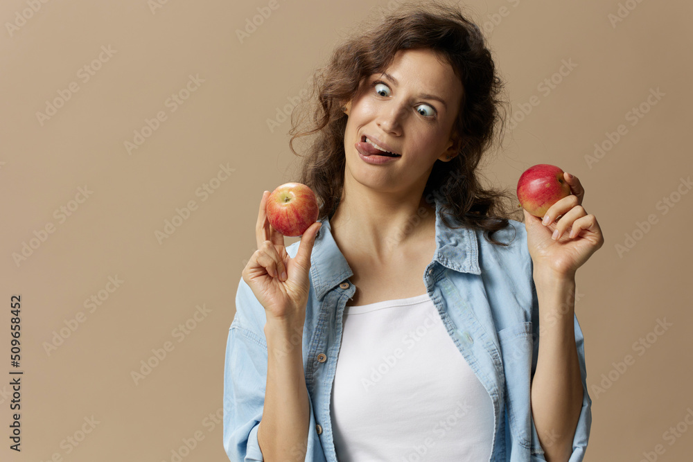 Crazy funny cute curly beautiful female in jeans casual shirt with apple squints eyes posing isolated on over beige pastel background. Healthy food. Natural eco-friendly products concept. Copy space Crazy funny cute curly beautiful female in jeans casual shirt with apple squints eyes posing isolated on over beige pastel background. Healthy food. Natural eco-friendly products concept. Copy space