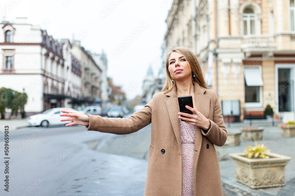Obraz premium Blonde woman catching car, holding smartphone with hand gesture in european city