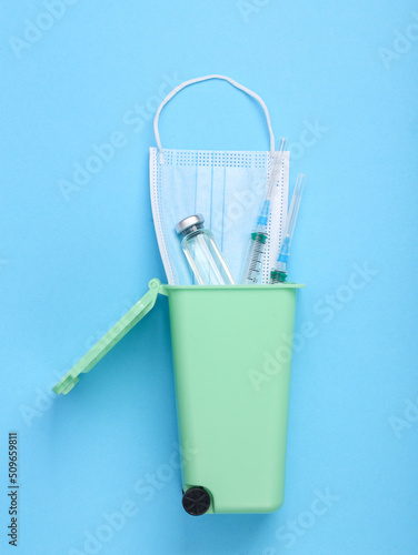 Vaccine bottle with syringe and medical mask in trash can on blue background