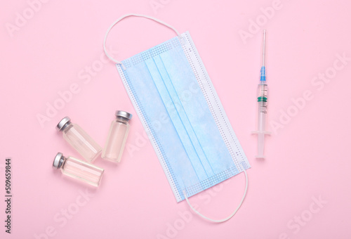 Vaccine bottle with a blank label for the brand and syringe, medical mask on pink background.