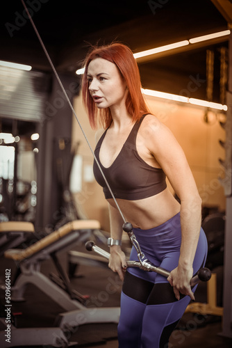 Red-haired young fitness woman in sportswear trains triceps muscles in a cable crossover exercise machine in a modern gym. Healthy lifestyle, fitness and bodybuilding concept
