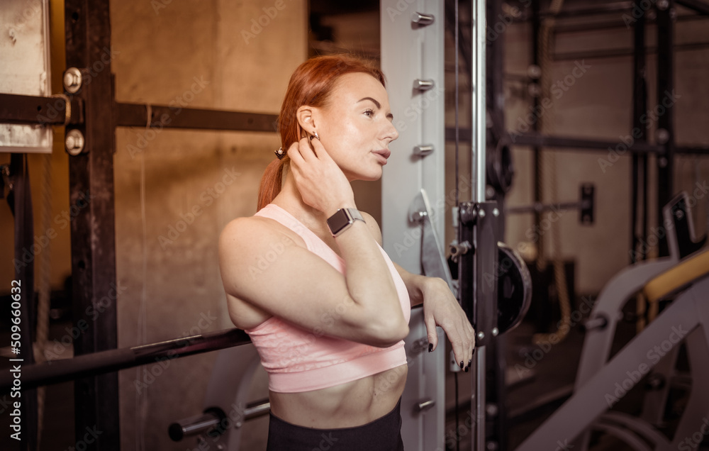 Fototapeta premium Attractive fit slim red-haired woman in sportswear resting leaning on barbell in modern gym