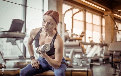Fit slim woman doing concentrated biceps dumbbell raises while sitting on a bench in a modern gym