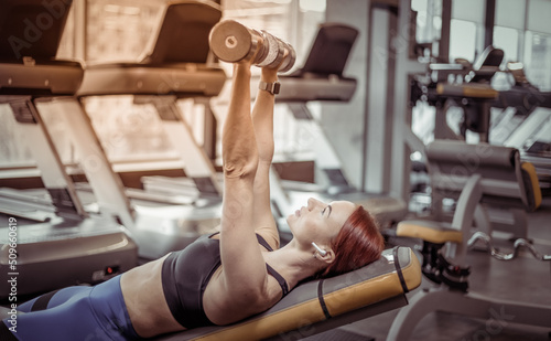 Fototapeta Naklejka Na Ścianę i Meble -  Athletic red-haired woman trains chest muscles with dumbbells in her hands while lying on an incline bench. Workout in the gym