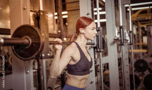 Portrait of athletic red-haired woman with barbell on shoulders in modern gym