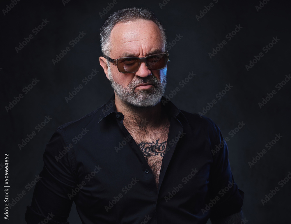 Studio shot of brutal aged macho with tattooed body dresed in black shirt and sunglasses.