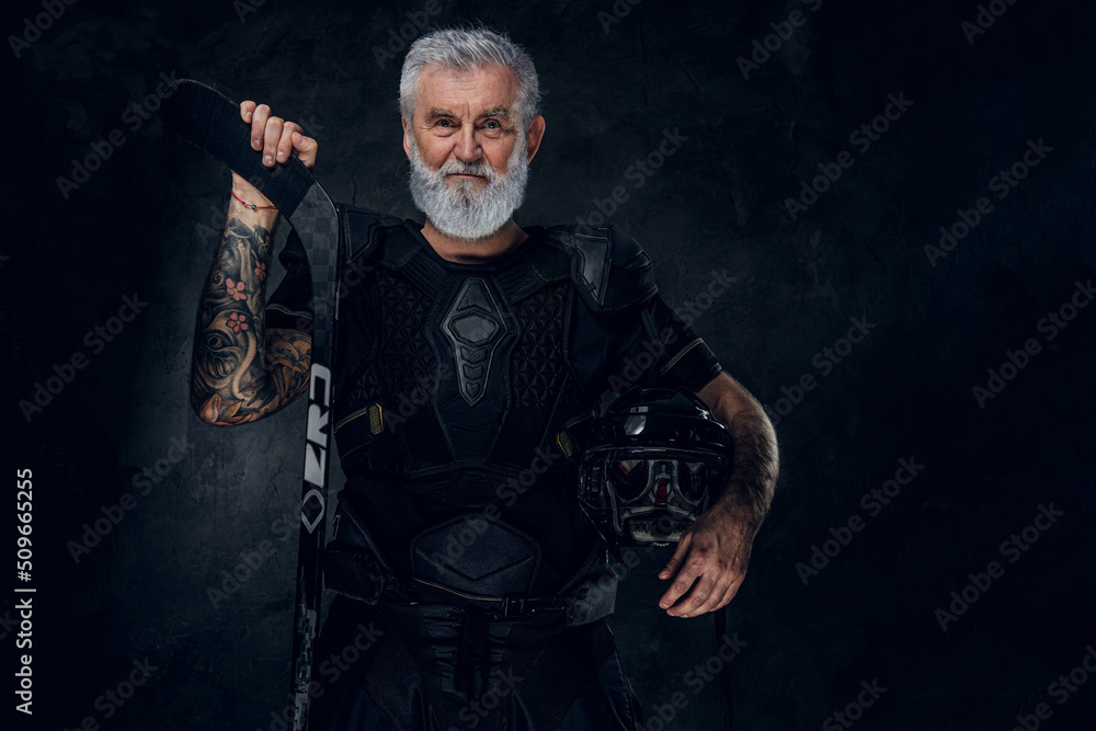 Shot of tattooed elderly hockey player with headwear and hockey stick against dark background.