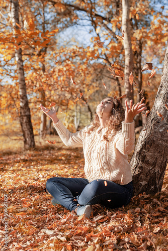 Young woman throwing yellow leaves up in the air sitting in the autumn forest