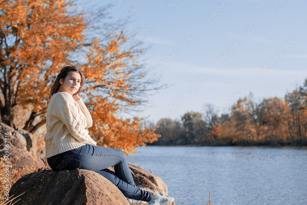 Thoughtful romantic woman sitting on rocks on the river bank in sunset in autumn day Thoughtful romantic woman sitting on rocks on the river bank in sunset in autumn day