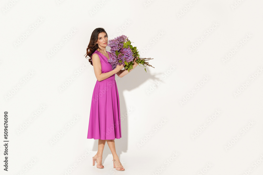 Full-length studio fashion portrait of elegant woman wearing purple dress and holding lilac brunches against white wall. International woman’s day, Mother’s day and spring concept. Full-length studio fashion portrait of elegant woman wearing purple dress and holding lilac brunches against white wall. International woman’s day, Mother’s day and spring concept.