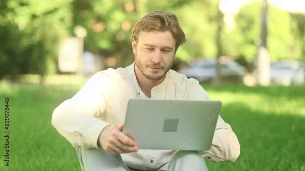 Portrait of handsome young man sitting on a grass with laptop and looking at the camera. Student learning online. Freelancer outside. Portrait of handsome young man sitting on a grass with laptop and looking at the camera. Student learning online. Freelancer outside.