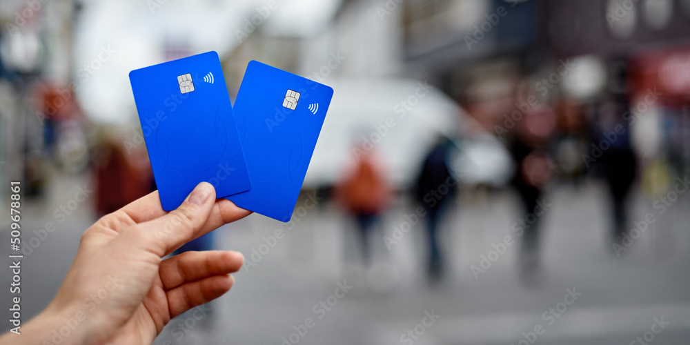 Fototapeta premium Two bank cards in the hand of a young girl against the background of a blurred street. Universal bank cards with chip and contactless payment.