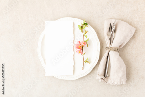 Festive or wedding table setting, porcelain empty plate, napking and cutlery with spring pink flowers on beige stone background. Floral elegant decor, top view, flat lay