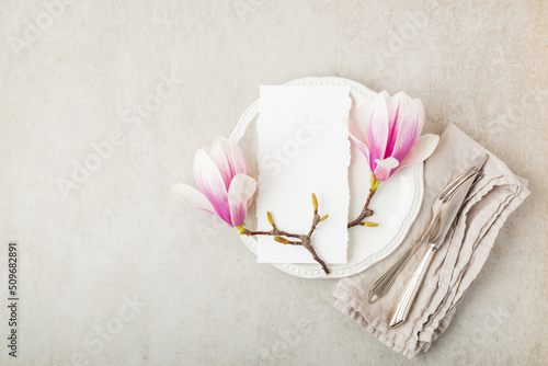 Festive or wedding table setting, porcelain empty plate, napking and cutlery with spring pink magnolia  flowers on beige stone background. Floral elegant decor, top view, flat lay photo