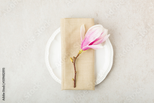 Festive or wedding table setting, porcelain empty plate, napking and cutlery with spring pink magnolia  flowers on beige stone background. Floral elegant decor, top view, flat lay
