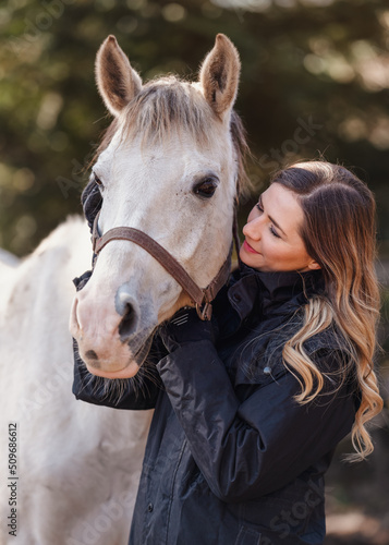 Young woman standing next to white Arabian horse, blurred trees background, closeup detail