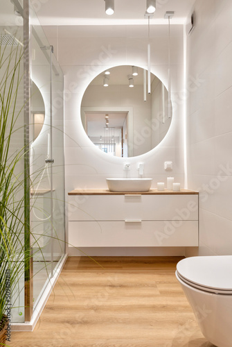 Luxury bathroom with glass to shower, round mirror with led lights, stylish washbasin and wooden floor. Modern interior of bathroom with wc and bath. Vertical.