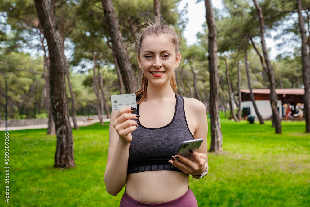 Cheerful brunette woman wearing sportive clothes on city park, outdoors showing plastic credit or debit card while holding mobile phone. Online shopping, banking and transaction concepts. Cheerful brunette woman wearing sportive clothes on city park, outdoors showing plastic credit or debit card while holding mobile phone. Online shopping, banking and transaction concepts.