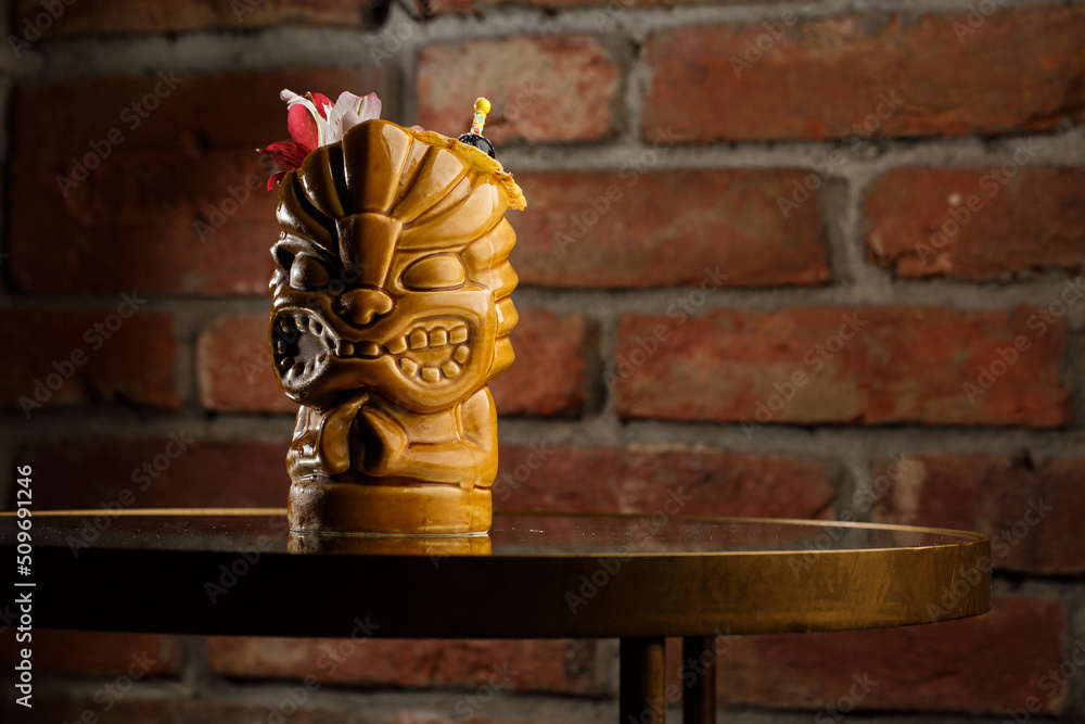Tropical cocktail - zombie, served in a tiki style with fruits and flowers. An exotic cocktail in a tiki glass stands on a wooden bar counter in a pub or restaurant, space for text.