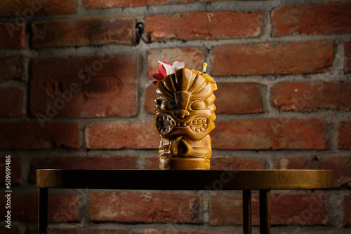 Obraz na płótnie Tropical cocktail - zombie, served in a tiki style with fruits and flowers. An exotic cocktail in a tiki glass stands on a wooden bar counter in a pub or restaurant, space for text. w salonie