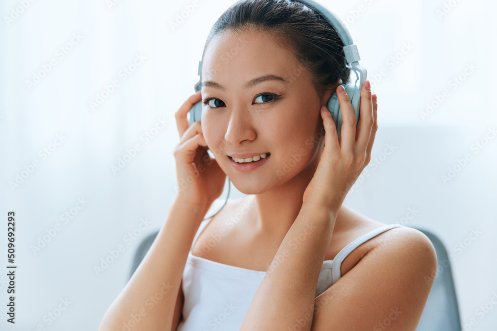 Closeup portrait of smiling happy tanned lovely young Asian woman in headphones looks at camera at home interior living room. Sound Studio Stream, Social media concept. Cool offer Banner Wide angle Closeup portrait of smiling happy tanned lovely young Asian woman in headphones looks at camera at home interior living room. Sound Studio Stream, Social media concept. Cool offer Banner Wide angle