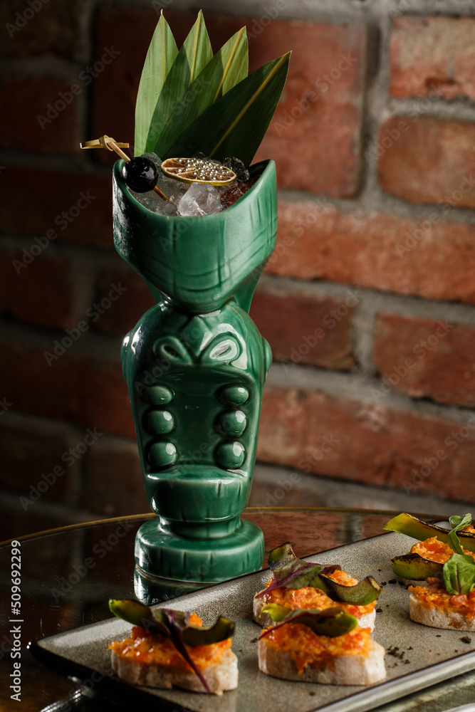 Tropical cocktail - zombie, served in a tiki style with fruits and flowers. An exotic cocktail in a tiki glass stands on a wooden bar counter in a pub or restaurant, space for text.