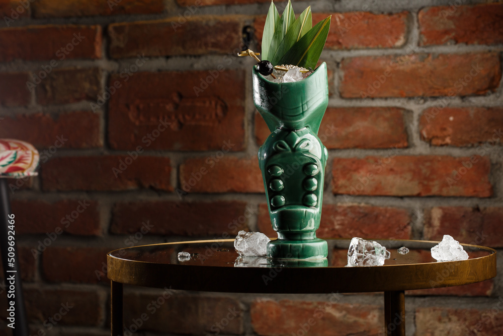 Tropical cocktail - zombie, served in a tiki style with fruits and flowers. An exotic cocktail in a tiki glass stands on a wooden bar counter in a pub or restaurant, space for text.