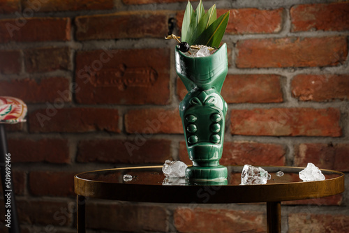 Obraz na płótnie Tropical cocktail - zombie, served in a tiki style with fruits and flowers. An exotic cocktail in a tiki glass stands on a wooden bar counter in a pub or restaurant, space for text. w salonie