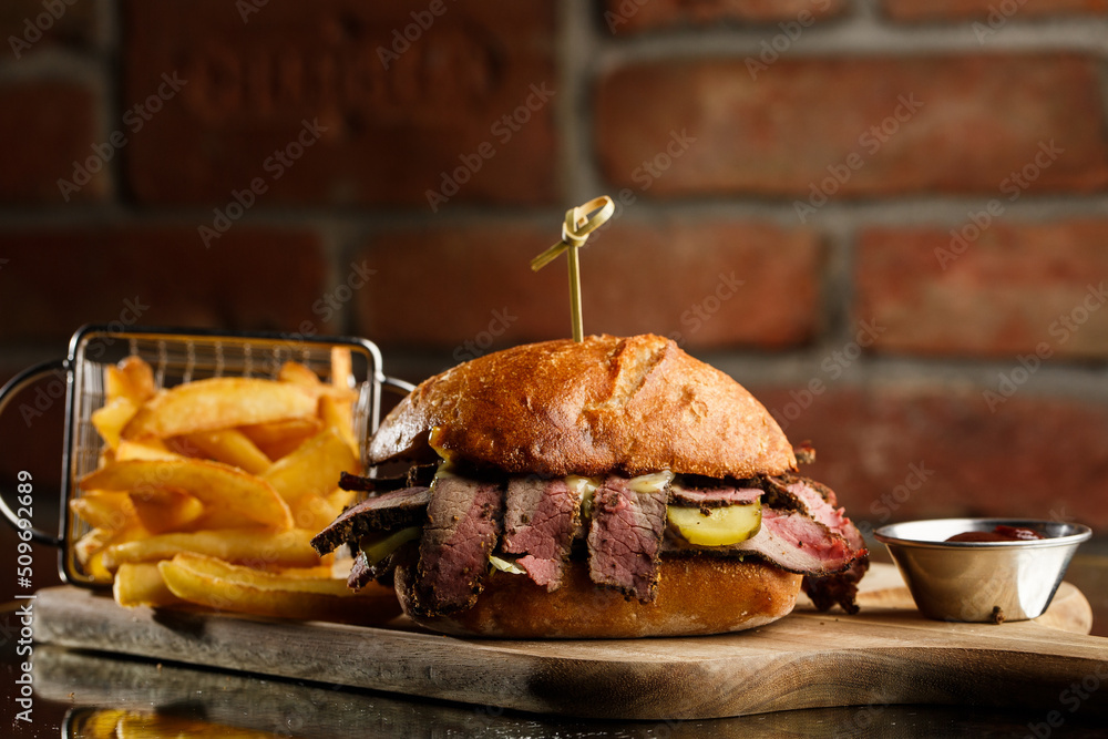 Pastrami sandwich on rye bread served with pickles and mustard sauce. Tasty sandwich with french fries potatoes on dark background.