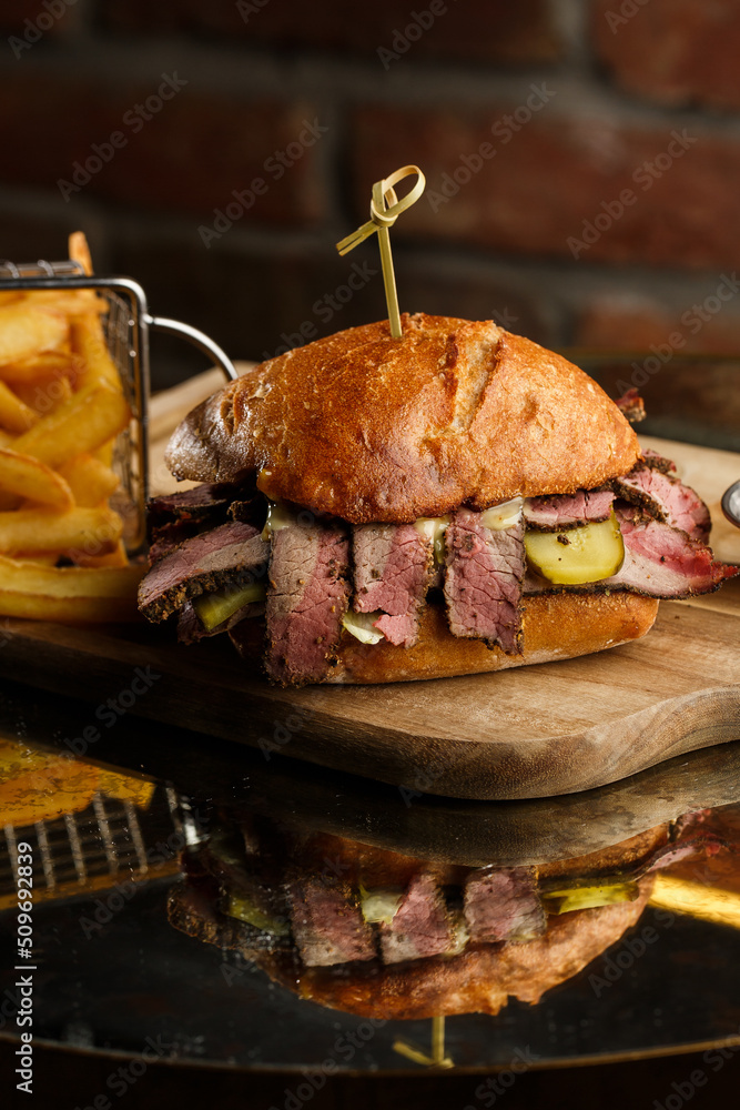 Pastrami sandwich on rye bread served with pickles and mustard sauce. Tasty sandwich with french fries potatoes on dark background.