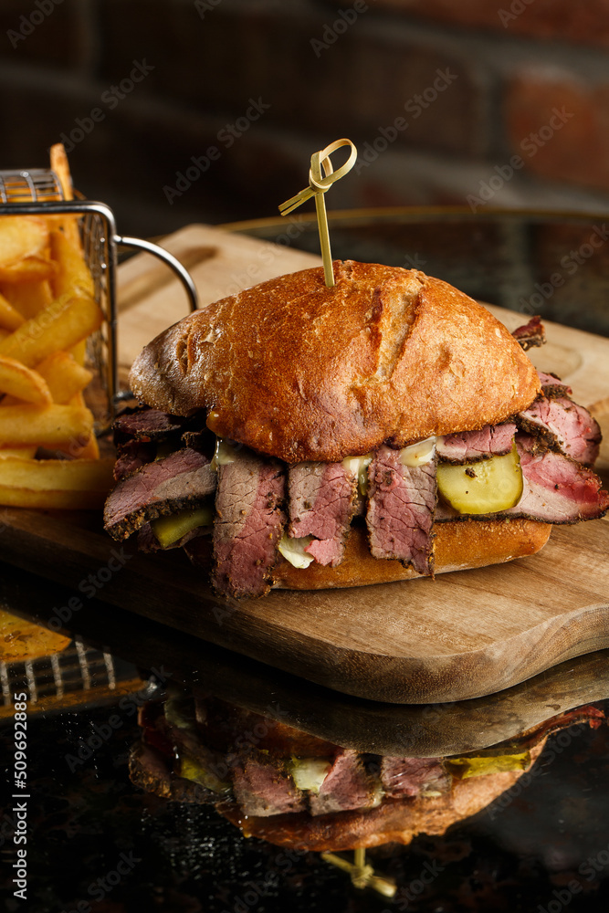 Pastrami sandwich on rye bread served with pickles and mustard sauce. Tasty sandwich with french fries potatoes on dark background.