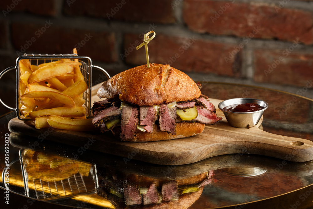 Pastrami sandwich on rye bread served with pickles and mustard sauce. Tasty sandwich with french fries potatoes on dark background.