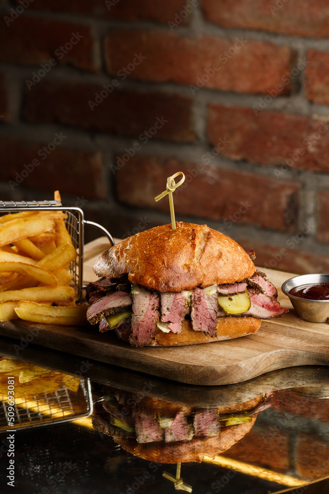 Pastrami sandwich on rye bread served with pickles and mustard sauce. Tasty sandwich with french fries potatoes on dark background.