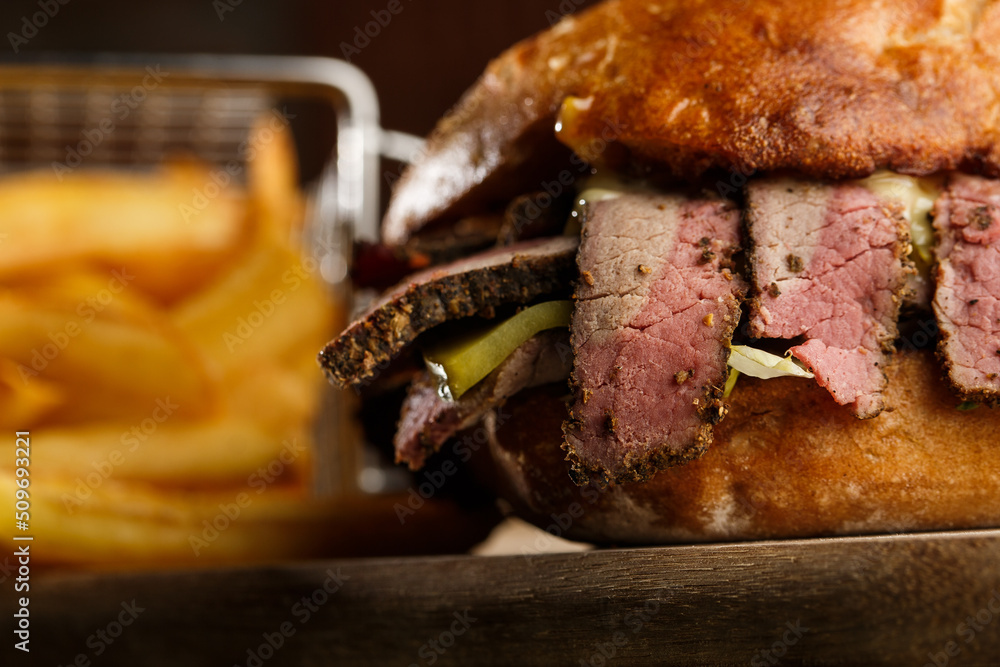 Pastrami sandwich on rye bread served with pickles and mustard sauce. Tasty sandwich with french fries potatoes on dark background.