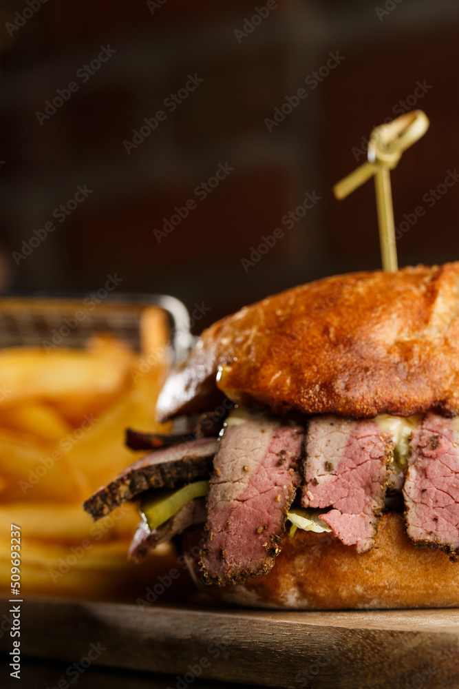 Pastrami sandwich on rye bread served with pickles and mustard sauce. Tasty sandwich with french fries potatoes on dark background.