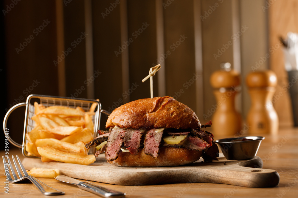 Pastrami sandwich on rye bread served with pickles and mustard sauce. Tasty sandwich with french fries potatoes on dark background.