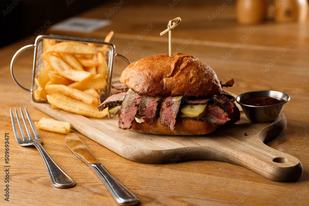 Pastrami sandwich on rye bread served with pickles and mustard sauce. Tasty sandwich with french fries potatoes on dark background.