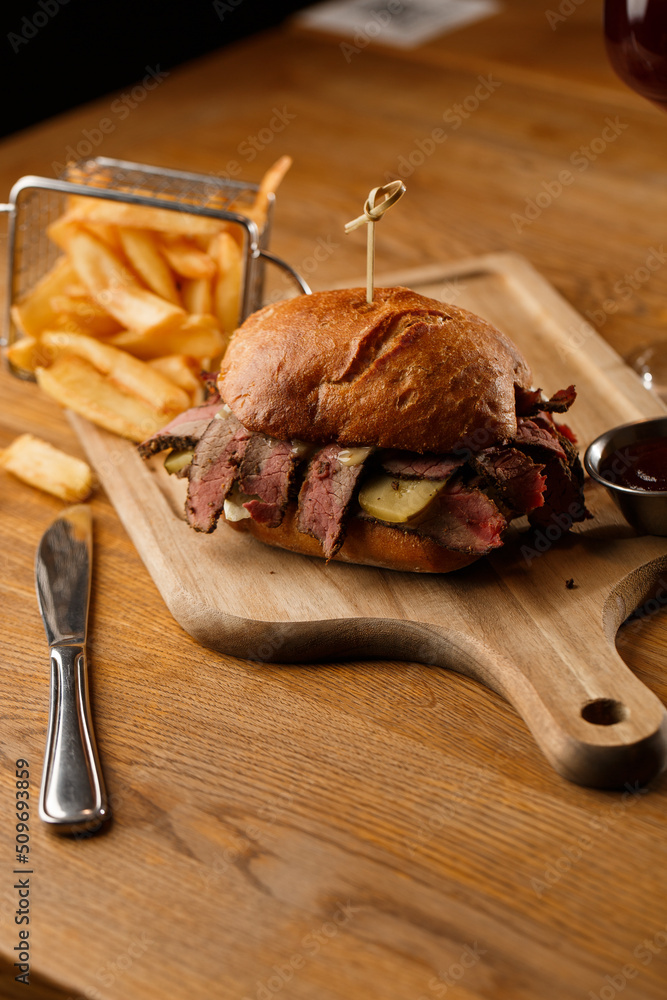 Pastrami sandwich on rye bread served with pickles and mustard sauce. Tasty sandwich with french fries potatoes on dark background.