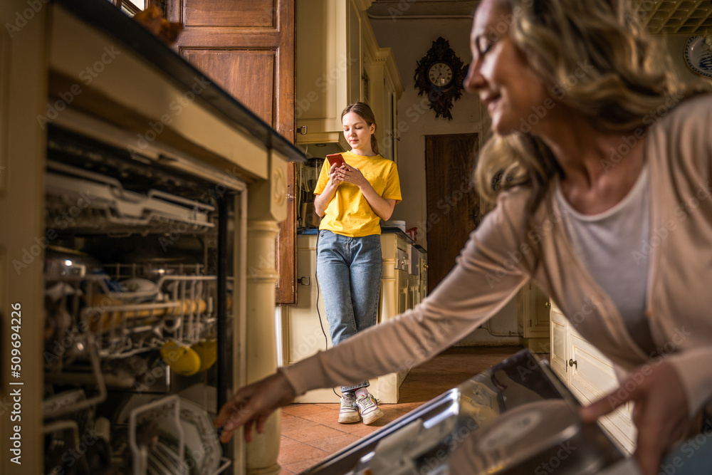 Calm woman put plates at the washing machine while her teen daughter reading messages Calm woman put plates at the washing machine while her teen daughter reading messages