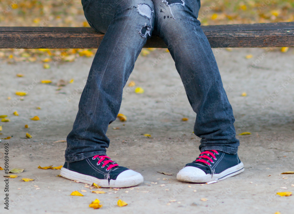 Legs of a teenage girl wearing sneakers and jeans sitting on the bench in the park in autumn Legs of a teenage girl wearing sneakers and jeans sitting on the bench in the park in autumn