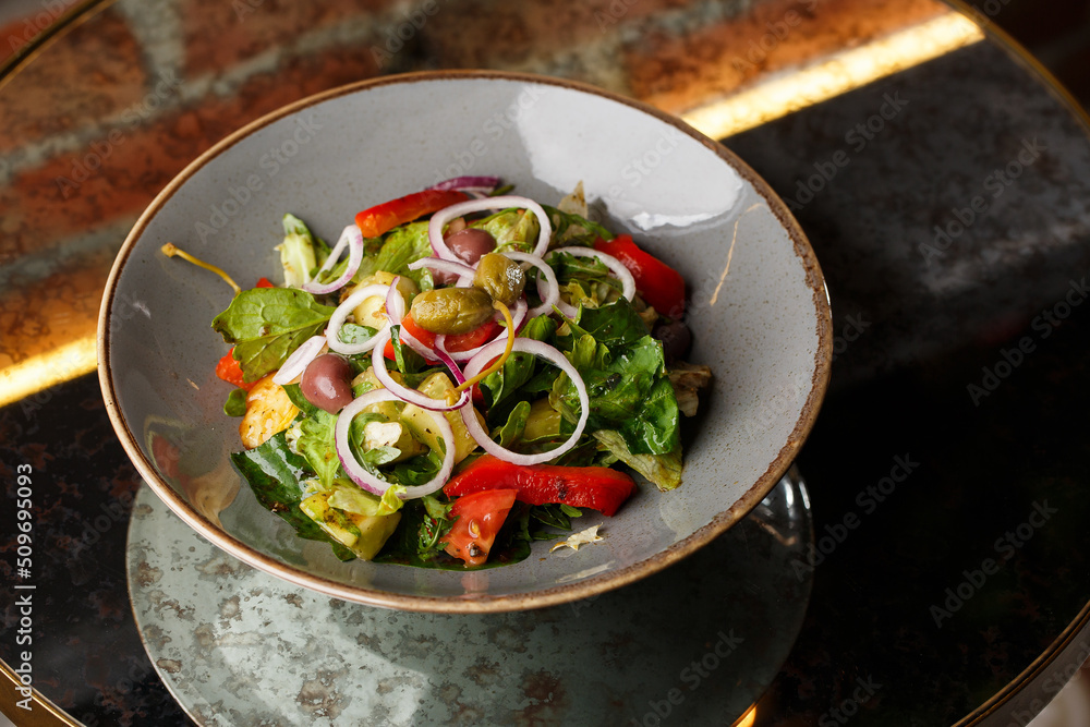 Greek salad with fresh vegetables: tomato, cucumber, red bel pepper, lettuce, onion, olives and cheese. Close-up on a white round plate on a wooden background. Salad menu with ingredients