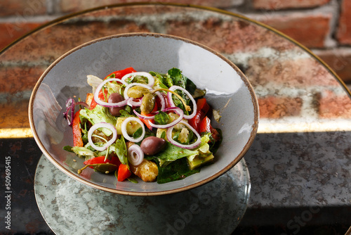 Greek salad with fresh vegetables: tomato, cucumber, red bel pepper, lettuce, onion, olives and cheese. Close-up on a white round plate on a wooden background. Salad menu with ingredients